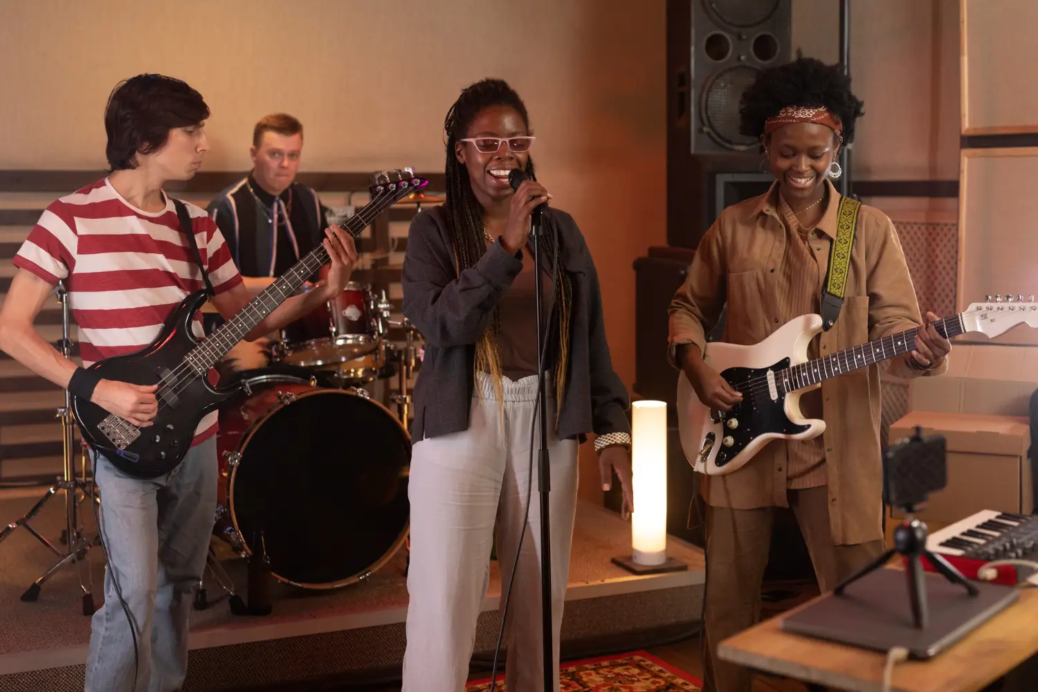 A four-member band rehearsing in a music studio. The group consists of a female vocalist standing center stage holding a wired microphone and smiling, a male bass guitarist on the left in a red and white striped t-shirt, a drummer in the background behind a red drum kit, and a female electric guitarist on the right in a brown outfit and red headband. Studio equipment, amplifiers, a lamp, and soundproofing panels are visible in the background. A microphone and laptop are positioned in the foreground on a table, suggesting possible recording or live streaming.