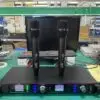 A Lindem Japan EA-10200 UHF professional wireless microphone system setup on a green workbench. The system includes two black handheld wireless microphones positioned vertically on top of the receiver unit. The dual-channel receiver displays frequency data (664.750 MHz and 665.150 MHz), battery status, and signal strength with multicolor LED indicators. A desktop monitor, tools, wiring, and testing equipment are visible in the background, suggesting a production or testing environment.