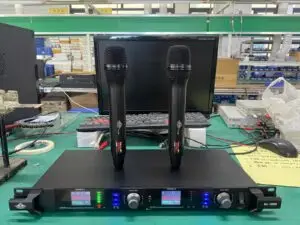 A Lindem Japan EA-10200 UHF professional wireless microphone system setup on a green workbench. The system includes two black handheld wireless microphones positioned vertically on top of the receiver unit. The dual-channel receiver displays frequency data (664.750 MHz and 665.150 MHz), battery status, and signal strength with multicolor LED indicators. A desktop monitor, tools, wiring, and testing equipment are visible in the background, suggesting a production or testing environment.
