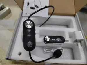 A wireless headset microphone system displayed in an open box with foam packaging. The main headset unit includes volume control buttons and a boom mic, labeled "DN-T1000" and "ETIMEK JAPAN." Also included are a compact receiver with a 3.5mm jack, a USB charging cable, a wall charger adapter, and an instruction manual in the background.