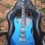 A metallic blue electric guitar with two humbucker pickups, a tremolo bridge, and a sharp double-cutaway body design, resting on a black gaming chair. The background features a brick-patterned wall with green moss accents.