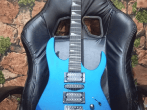 A metallic blue electric guitar with two humbucker pickups, a tremolo bridge, and a sharp double-cutaway body design, resting on a black gaming chair. The background features a brick-patterned wall with green moss accents.