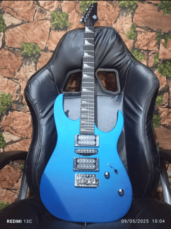 A metallic blue electric guitar with two humbucker pickups, a tremolo bridge, and a sharp double-cutaway body design, resting on a black gaming chair. The background features a brick-patterned wall with green moss accents.