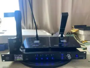 A professional UHF wireless conference microphone system setup, featuring a central receiver unit with LED indicators and knobs, two gooseneck-style desktop microphones with square bases, and a wireless handheld mic with a bodypack transmitter, all placed on a table with a curtain backdrop.