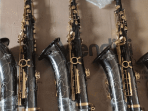 Four black alto saxophones with gold keys and floral engravings are arranged side by side on a cardboard surface. The instruments are partially wrapped in protective plastic, showcasing their polished finish and decorative craftsmanship, likely in a production or storage setting.
