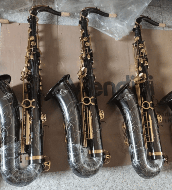 Four black alto saxophones with gold keys and floral engravings are arranged side by side on a cardboard surface. The instruments are partially wrapped in protective plastic, showcasing their polished finish and decorative craftsmanship, likely in a production or storage setting.