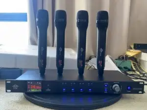 A Lindem Japan EA-10400 UHF four-channel wireless microphone system displayed on a round surface in front of a curtained window. The setup features four identical black handheld microphones standing upright on the receiver unit. The front panel of the receiver shows frequency data (e.g., 648.000 MHz), LED indicators for RF and AF signals for each channel, a manual frequency dial, IR sync, and power button. Documents, tools, and packaging are visible in the background, indicating a testing or assembly environment.