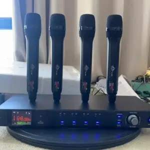 A Lindem Japan EA-10400 UHF four-channel wireless microphone system displayed on a round surface in front of a curtained window. The setup features four identical black handheld microphones standing upright on the receiver unit. The front panel of the receiver shows frequency data (e.g., 648.000 MHz), LED indicators for RF and AF signals for each channel, a manual frequency dial, IR sync, and power button. Documents, tools, and packaging are visible in the background, indicating a testing or assembly environment.