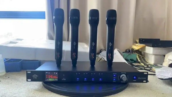 A Lindem Japan EA-10400 UHF four-channel wireless microphone system displayed on a round surface in front of a curtained window. The setup features four identical black handheld microphones standing upright on the receiver unit. The front panel of the receiver shows frequency data (e.g., 648.000 MHz), LED indicators for RF and AF signals for each channel, a manual frequency dial, IR sync, and power button. Documents, tools, and packaging are visible in the background, indicating a testing or assembly environment.