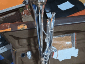 A shiny silver trumpet placed inside a brown velvet-lined protective case. The case also contains a white glove, a small cleaning cloth in a plastic pouch, a plastic bottle of valve oil, and a mouthpiece. A black office chair and part of a music studio are visible in the background.