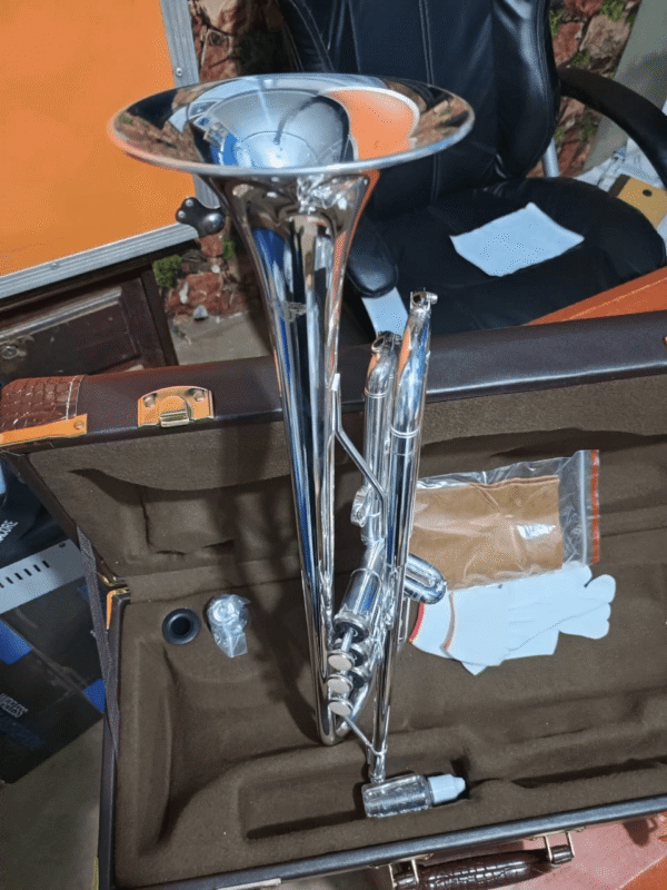 A shiny silver trumpet placed inside a brown velvet-lined protective case. The case also contains a white glove, a small cleaning cloth in a plastic pouch, a plastic bottle of valve oil, and a mouthpiece. A black office chair and part of a music studio are visible in the background.