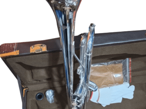 A silver trumpet resting in an open brown case lined with soft material. Inside the case, there are trumpet accessories including a pair of white gloves, a small bottle of valve oil, and a mouthpiece in plastic wrapping.