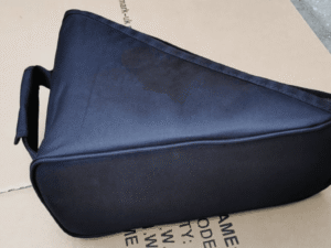 A black triangular padded carrying case resting on a cardboard surface outdoors. The case has a sturdy handle on one side and appears designed to protect and transport equipment or instruments, made from durable fabric material.