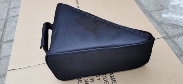 A black triangular padded carrying case resting on a cardboard surface outdoors. The case has a sturdy handle on one side and appears designed to protect and transport equipment or instruments, made from durable fabric material.