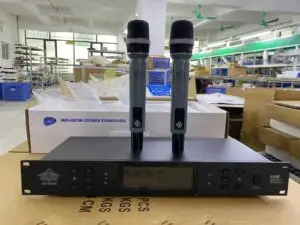 A dual UHF wireless microphone system featuring two handheld microphones placed vertically on top of a receiver unit. The unit has a digital display, control buttons, and is labeled EA-8200C by EIMEX JAPAN. The setup is photographed in an electronics manufacturing facility with boxes, shelves, and packing materials visible in the background.