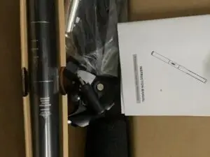 An open cardboard box containing a professional condenser microphone kit. Inside, there is a long black condenser microphone, a coiled cable wrapped in plastic, a foam windscreen, a microphone clip, and an instruction manual. The items are neatly arranged in separate compartments within the box.