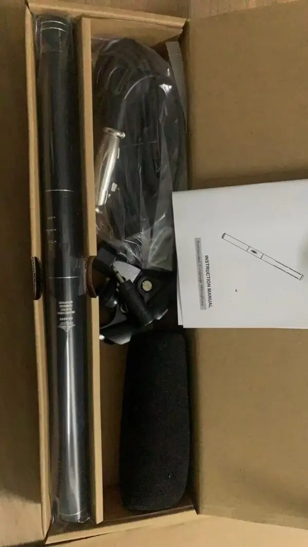 An open cardboard box containing a professional condenser microphone kit. Inside, there is a long black condenser microphone, a coiled cable wrapped in plastic, a foam windscreen, a microphone clip, and an instruction manual. The items are neatly arranged in separate compartments within the box.