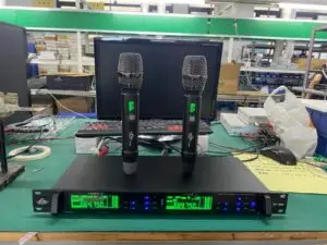 A UHF professional wireless microphone system model EA-9900 displayed on a workbench. Two handheld wireless microphones are standing on top of the receiver unit. The receiver's digital display is illuminated in green, showing channel frequencies (664.750 MHz and 689.750 MHz), signal strength, and volume levels. The setting appears to be a manufacturing or testing facility with a keyboard, monitor, and various equipment in the background.