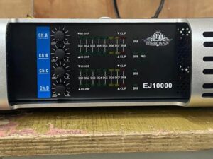A silver and black Ejimek Japan EJ10000 audio amplifier resting on a worn wooden surface. The front panel displays four blue channel volume knobs labeled Ch.A, Ch.B, Ch.C, and Ch.D, alongside LED signal meters and clip indicators. The amplifier has ventilation slots on both sides.