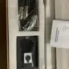 An open cardboard box containing a professional condenser microphone kit. Inside, there is a long black condenser microphone, a coiled cable wrapped in plastic, a foam windscreen, a microphone clip, and an instruction manual. The items are neatly arranged in separate compartments within the box.