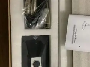 An open cardboard box containing a professional condenser microphone kit. Inside, there is a long black condenser microphone, a coiled cable wrapped in plastic, a foam windscreen, a microphone clip, and an instruction manual. The items are neatly arranged in separate compartments within the box.