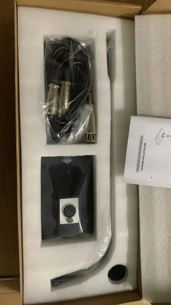 An open cardboard box containing a professional condenser microphone kit. Inside, there is a long black condenser microphone, a coiled cable wrapped in plastic, a foam windscreen, a microphone clip, and an instruction manual. The items are neatly arranged in separate compartments within the box.