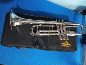 A polished silver trumpet resting on a black soft carrying case with the gold "Ejimek Japan" logo. The case is placed on a bright blue carpeted surface, and the trumpet is shown from the side, highlighting the valves and bell.