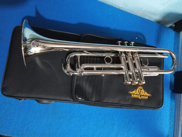 A polished silver trumpet resting on a black soft carrying case with the gold "Ejimek Japan" logo. The case is placed on a bright blue carpeted surface, and the trumpet is shown from the side, highlighting the valves and bell.