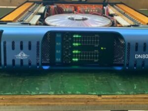 A blue Ejimek Japan DN8000 audio amplifier placed on a green wooden surface. The front panel features four volume knobs with LED signal meters and indicators. The top cover of the amplifier is removed, revealing internal components including circuit boards and a large cooling fan.