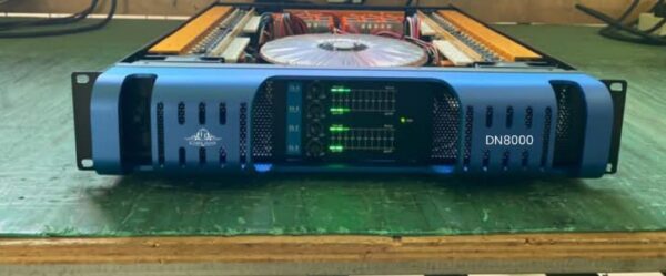 A blue Ejimek Japan DN8000 audio amplifier placed on a green wooden surface. The front panel features four volume knobs with LED signal meters and indicators. The top cover of the amplifier is removed, revealing internal components including circuit boards and a large cooling fan.