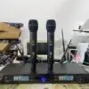 A Lindemaier EA-6000 UHF wireless microphone system on a workbench. Two black handheld microphones are standing upright on top of the receiver unit. The receiver's dual-channel display shows operating frequencies (650.250 MHz and 686.750 MHz), signal levels, and other telemetry. The setup is surrounded by electronic testing equipment, cables, and boxes, indicating a testing or assembly environment.