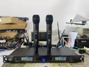 A Lindemaier EA-6000 UHF wireless microphone system on a workbench. Two black handheld microphones are standing upright on top of the receiver unit. The receiver's dual-channel display shows operating frequencies (650.250 MHz and 686.750 MHz), signal levels, and other telemetry. The setup is surrounded by electronic testing equipment, cables, and boxes, indicating a testing or assembly environment.