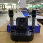 A dual wireless microphone system featuring two handheld microphones placed in a black charging dock with a digital display labeled "UHF Professional Wireless Microphone EA-7200C." The setup is positioned on a cardboard surface in an industrial or assembly workspace.