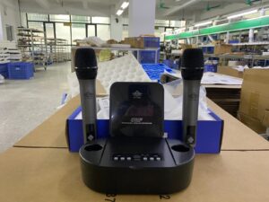 A dual wireless microphone system featuring two handheld microphones placed in a black charging dock with a digital display labeled "UHF Professional Wireless Microphone EA-7200C." The setup is positioned on a cardboard surface in an industrial or assembly workspace.