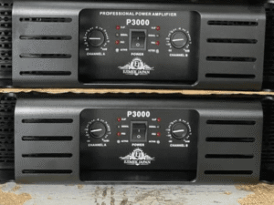 Two black Ejimek Japan professional power amplifiers model P3000 stacked on top of each other. Each amplifier has a front panel with two control knobs labeled Channel A and Channel B, a power switch, and LED indicators for status such as clip, signal, and active. The amplifiers are placed on a rough surface with a cardboard divider between them.
