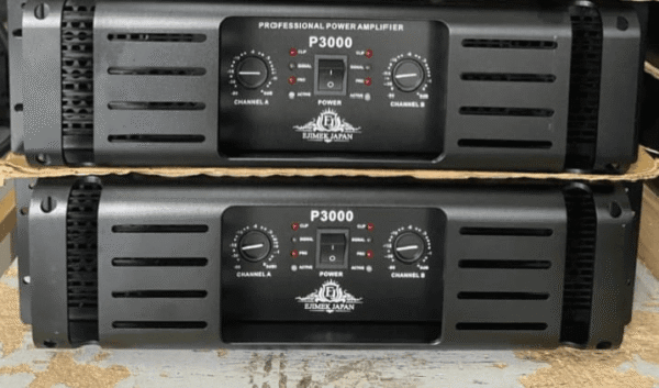 Two black Ejimek Japan professional power amplifiers model P3000 stacked on top of each other. Each amplifier has a front panel with two control knobs labeled Channel A and Channel B, a power switch, and LED indicators for status such as clip, signal, and active. The amplifiers are placed on a rough surface with a cardboard divider between them.