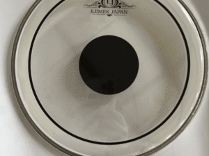 Clear drum head featuring a black center dot and the "EJIMEK JAPAN" logo printed near the top. The drum head is placed on a white surface with visible shadows and light reflections.