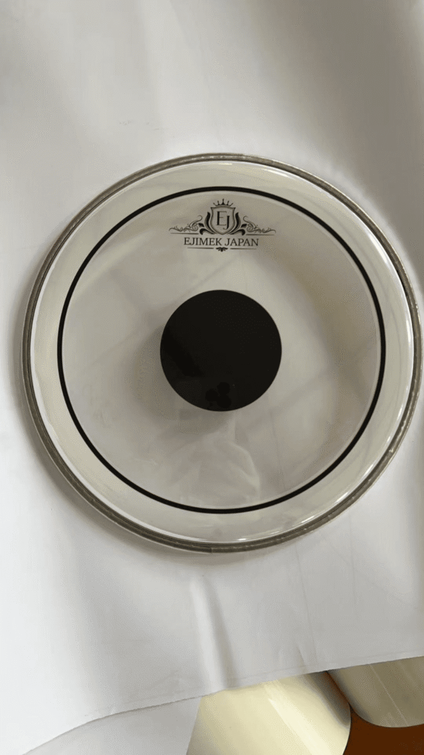 Clear drum head featuring a black center dot and the "EJIMEK JAPAN" logo printed near the top. The drum head is placed on a white surface with visible shadows and light reflections.