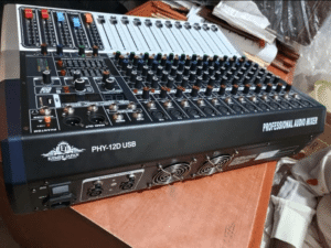 Ejimek Japan PHY-12D USB professional audio mixer placed on a wooden table. The mixer has multiple sliders, knobs, input ports, and control buttons. The device is black with white and colored controls, labeled “PROFESSIONAL AUDIO MIXER” on the side.