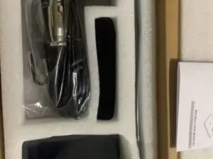 A set of microphone accessories neatly packed in a white foam insert inside a box. Visible items include an XLR cable wrapped in plastic, a black power adapter with a power button, a microphone stand rod, and a folded instruction manual. All components are organized in separate compartments.