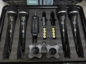 A professional wireless microphone kit featuring four black DN-W1500PRO UHF handheld microphones, a single wireless receiver with a 6.35mm plug, eight AA batteries, two anti-roll mic holders, a USB charging cable, and an instruction manual, all organized in a foam-lined black carrying case.