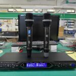 A professional UHF wireless microphone system featuring two handheld microphones standing on a digital receiver unit. The receiver displays channel information on two blue LCD screens. The setup is placed on a workbench in an electronics or manufacturing environment, with a computer and testing equipment in the background.