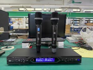 A professional UHF wireless microphone system featuring two handheld microphones standing on a digital receiver unit. The receiver displays channel information on two blue LCD screens. The setup is placed on a workbench in an electronics or manufacturing environment, with a computer and testing equipment in the background.