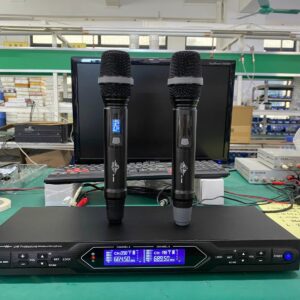 A professional UHF wireless microphone system featuring two handheld microphones standing on a digital receiver unit. The receiver displays channel information on two blue LCD screens. The setup is placed on a workbench in an electronics or manufacturing environment, with a computer and testing equipment in the background.