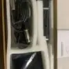 An open box containing a professional condenser microphone kit. The kit includes a coiled cable with XLR connectors, a black microphone body, a small black power box with an XLR input, and an instruction manual. All components are wrapped in plastic and snugly placed in white foam packaging inside the box.