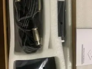 An open box containing a professional condenser microphone kit. The kit includes a coiled cable with XLR connectors, a black microphone body, a small black power box with an XLR input, and an instruction manual. All components are wrapped in plastic and snugly placed in white foam packaging inside the box.