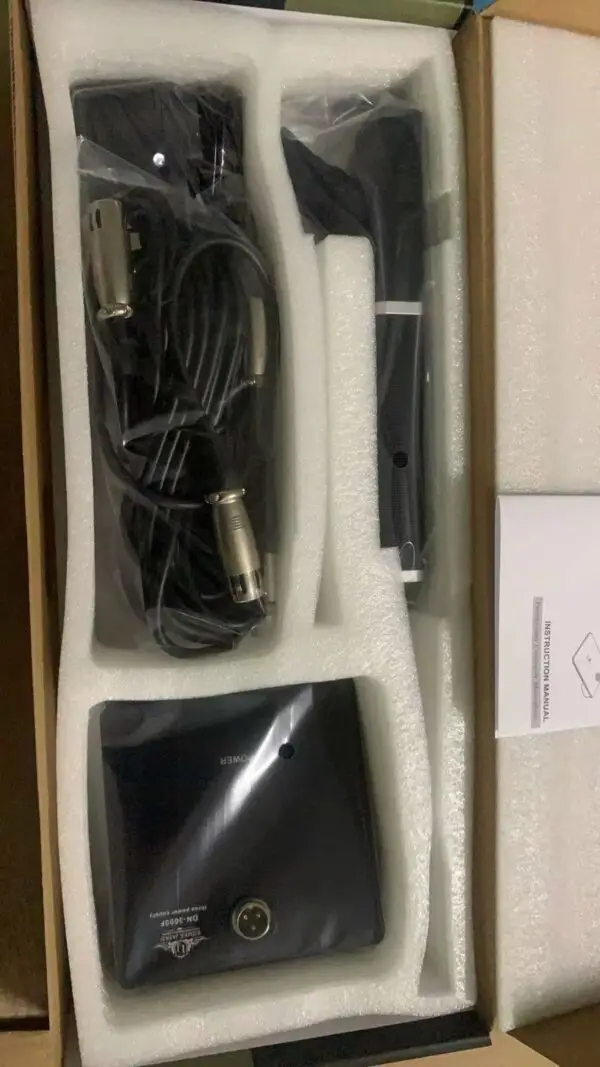 An open box containing a professional condenser microphone kit. The kit includes a coiled cable with XLR connectors, a black microphone body, a small black power box with an XLR input, and an instruction manual. All components are wrapped in plastic and snugly placed in white foam packaging inside the box.