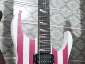 An electric guitar featuring a bold American flag design with red and white stripes and a blue star field on the body. The guitar has a black headstock, black hardware, two humbucker pickups, and is mounted vertically on a patterned wall.