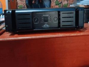 A black Ejimek Japan professional power amplifier model P4000 sits on a wooden desk. The front panel features two control knobs labeled Channel A and Channel B, a power switch, and LED indicators for active, power, and other statuses. In the background is a black leather chair and a wall with a stone pattern.
